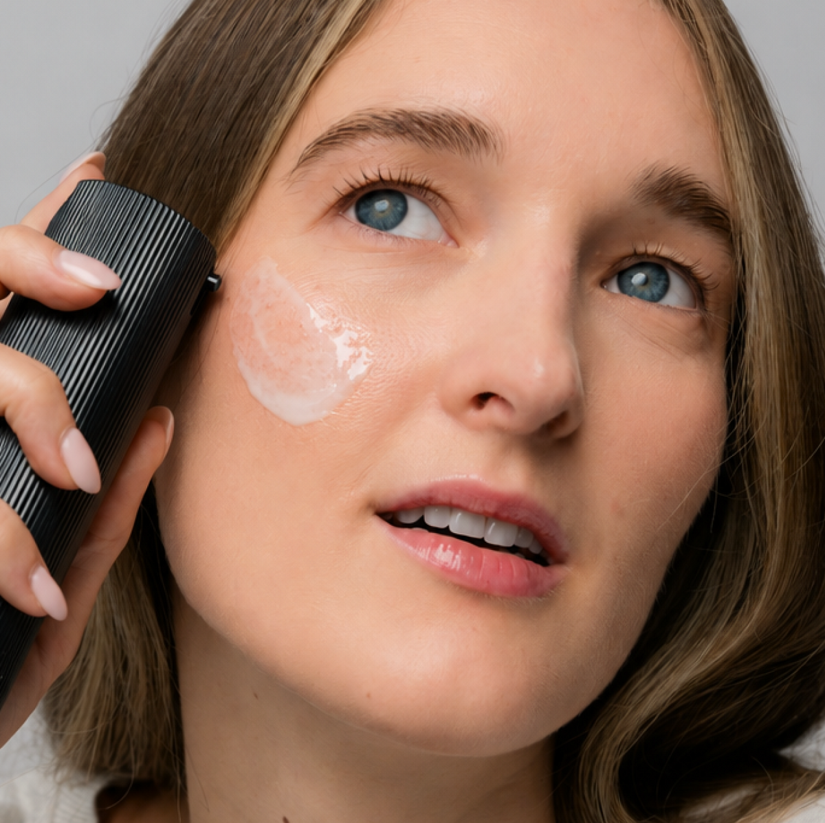 A model holding the Facial Serum bottle to her cheek, with a swatch of white cream on her skin.