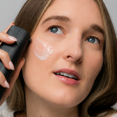 A model holding the Facial Serum bottle to her cheek, with a swatch of white cream on her skin.